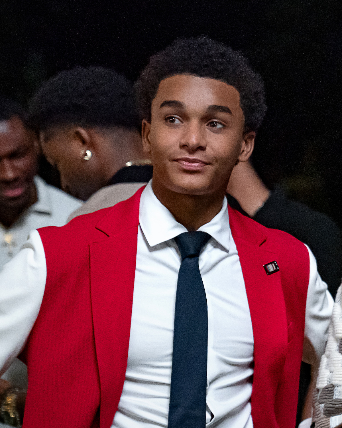Young man in formal attire with a red blazer, white shirt, and dark tie standing at an indoor event. Other people in formalwear are in the background, the setting appears social and lively.