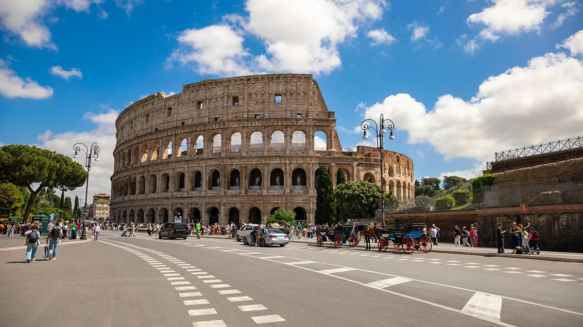 A stunning panoramic view of the iconic Colosseum in Rome, Italy, captured on a bright sunny day with tourists, city traffic, and horse-drawn carriages bringing life to the historic surroundings. The image blends Ancient Roman heritage with contemporary urban movement, showcasing one of Europe’s most famous travel destinations under dramatic blue skies and soft clouds.