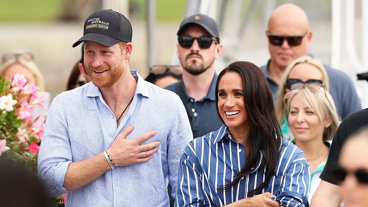 Prince Harry and Meghan Markle in casual clothes smiling and waving to a crowd in Australia.